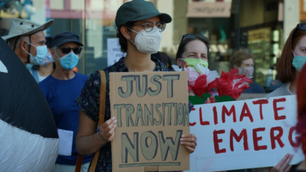 A young person wearing an N95 mask with a maple leaf on it holds a sign that reads "Just Transition Now"