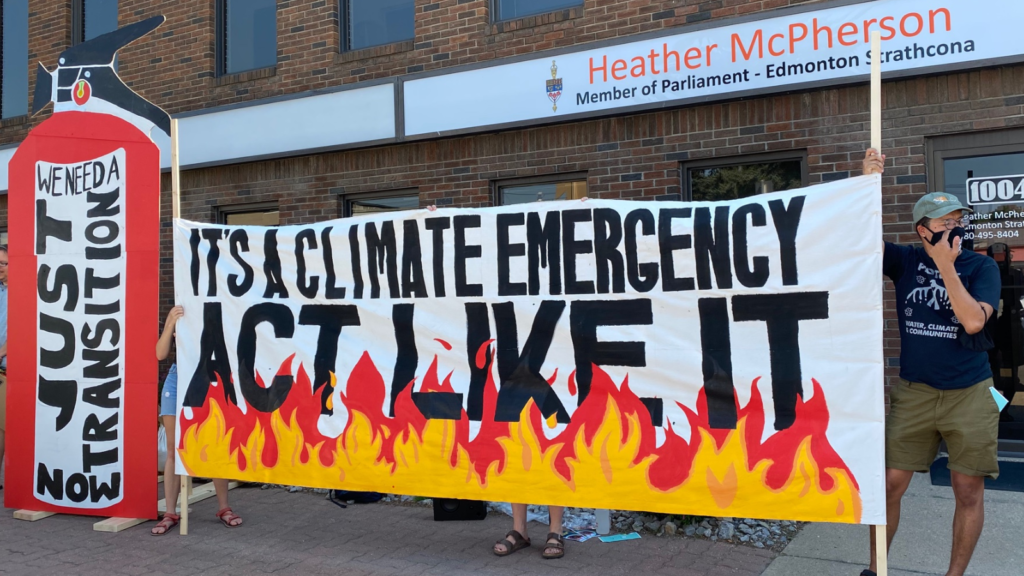 Concerned citizens stand outside Edmonton MP Heather McPherson's office holding a giant banner that reads "It's a climate emergency. Act like it". They also have a giant fire extinguisher with text on it that says "we need a just transition now"