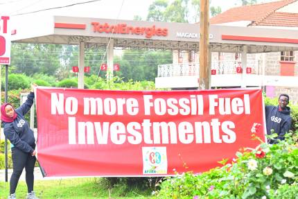 Aktivist*innen halten einen Banner auf dem "No more Fossil Fuel Investments" steht. Im Hintergrund ist eine Tankstelle von TOTAL zu sehen.