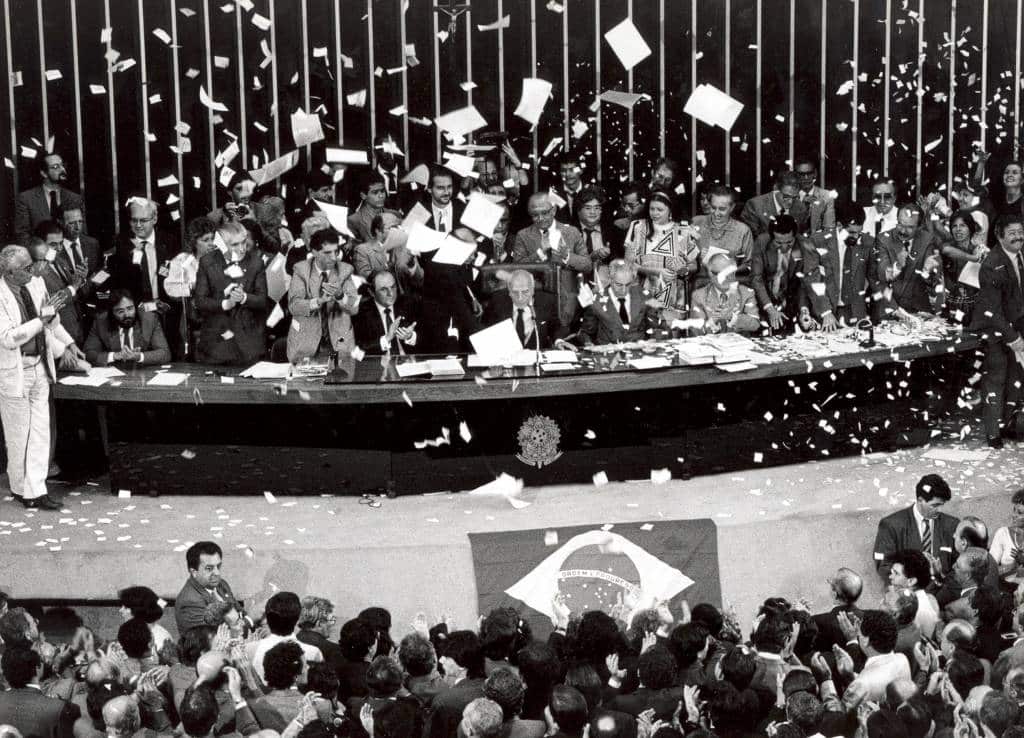 Imagem em preto e branco, Ulysses Guimarães com parlamentares festejam em meio a papéis picados. pronunciou as palavras históricas: "Declaro promulgado o documento da liberdade, da democracia e da justiça social do Brasil"