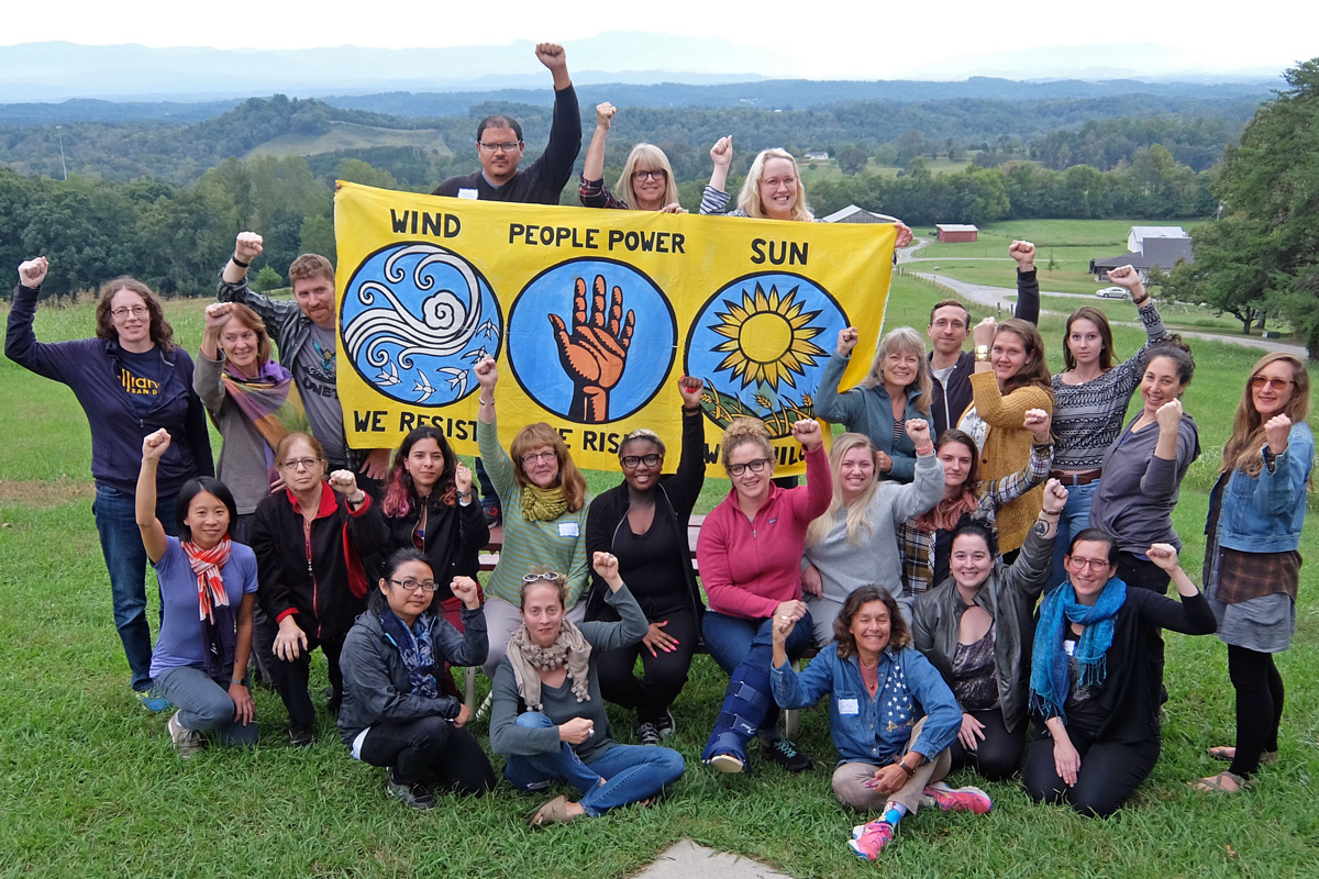 Group on a grassy hill with fists in the air holding a banner that represents the power of wind, sun, and people rising together