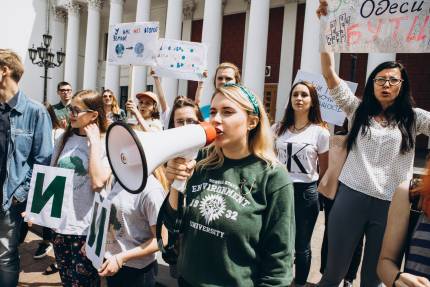 Young woman in green sweater holding megaphone and speaking to crowd outside