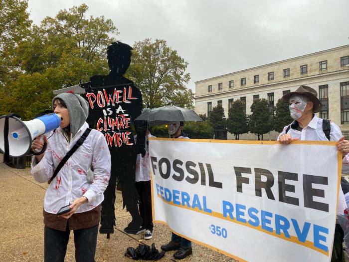 In the rain outside the DC Federal Reserve building, A cutout of a person that says "Powell is a Climate Zombie," next to two people holding a banner reading "Fossil Free Federal Reserve" behind a person speaking into a megaphone wearing a Zombie costume.