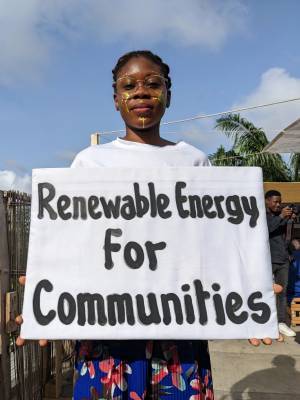 A woman is facing the camera, holding a sign saying 'renewable energy for communities'