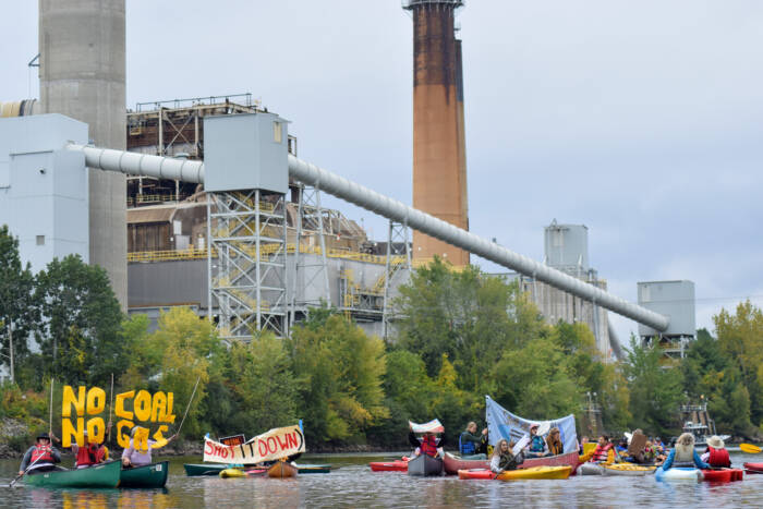 Climate activists sit in kayaks on a river holding up signs that say "No Coal No Gas" and "Shut it Down." They are in front of a coal plant.
