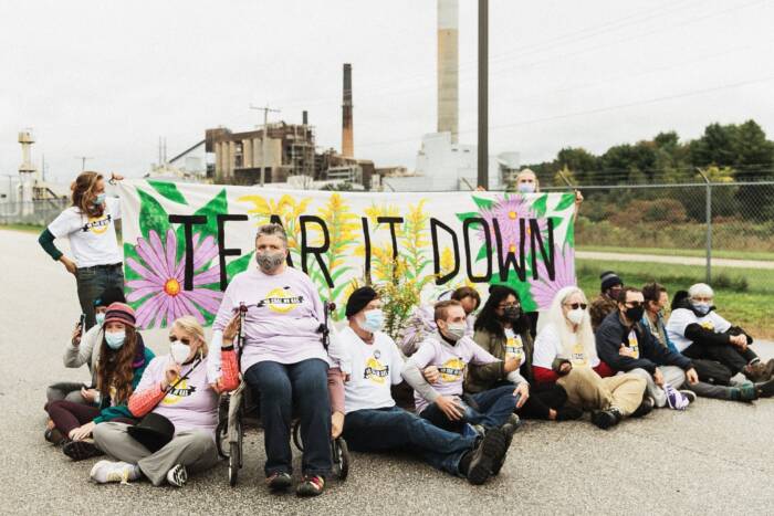 Activists from 350NH sitting and standing in front of banner that says "tear it down." In the background is the Merrimack station coal plant. 