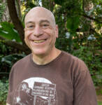 This photo shows Jeff Ordower, a white middle-aged man wearing a brown shirt. Jeff is smiling and standing outside in front of some trees. 