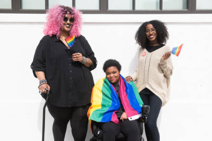 Three Black and disabled folx decked out happily in rainbow pride flags. On the left, a non-binary person holds a cane in one hand and waves a mini flag in the other. In the middle, a non-binary person sits in their power wheelchair, draped in a large flag. On the right, a femme rests one hand on their friend's shoulder and waves a mini flag around with the other hand. CC: Disabled and Here