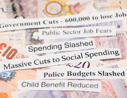 A background of money with ripped paper headlines on top. Headlines read spending slashed, massive cuts to social spending, child benefit reduced, etc.